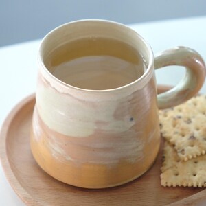 May include: A handmade ceramic mug with a beige and orange glaze, filled with a light-colored beverage, rests on a small wooden plate. Two crackers with seeds are placed next to the mug. The mug has a rounded handle.