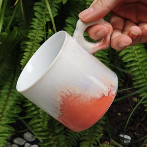 May include: White ceramic mug with a unique handle, featuring a gradient of colors from white to orange. The mug is held against a backdrop of green foliage, showcasing its design and shape.