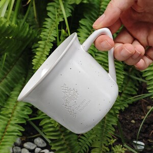 May include: A white speckled ceramic mug with a curved handle. The mug has an embossed design and the text "Yu Garden Shanghai". The mug is held in a hand against a background of green foliage and small rocks.