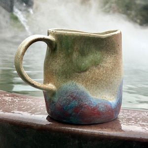 May include: A handcrafted ceramic mug with an irregular form. The mug displays a mix of colours: green, tan, blue, and red, producing a textured, watercolour-like appearance. It has a robust handle and is set against a blurred backdrop of water and steam.