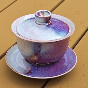 May include: A ceramic tea set with a purple, blue, and white glaze. The set includes a lidded bowl and a saucer. The lid has a small handle. The tea set is designed for serving tea.