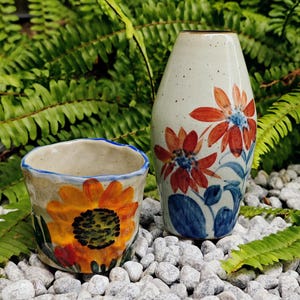 May include: Two ceramic vases with floral designs. One is a small cup with a sunflower, the other is a tall vase with red and blue flowers. The vases are on a bed of white stones, with green foliage in the background.