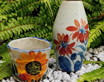 May include: Two ceramic vases with floral designs. One is a small cup with a sunflower, the other is a tall vase with red and blue flowers. The vases are on a bed of white stones, with green foliage in the background.