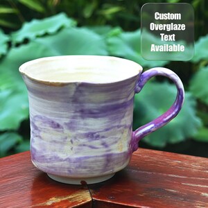 May include: A handmade ceramic mug with a white interior and a purple and white marbled exterior. The handle is purple. The mug has the text "Custom Overglaze Text Available".