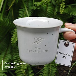 May include: A speckled white ceramic mug with a heart and arrow design, text that reads "Your Text / Logo Here," and a tag that says "Personalize Me!" The mug has a handle and is set against a green leafy background. The text "Custom Engraving Available!" is also visible.