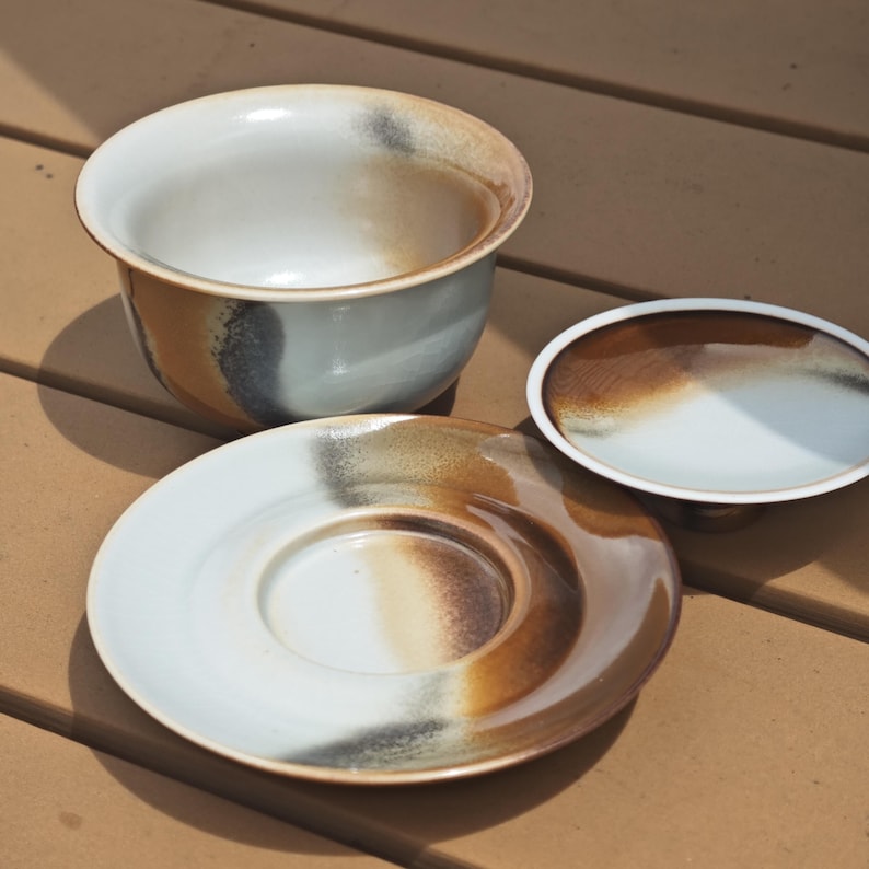May include: A ceramic tea set consisting of a bowl, a saucer, and a small plate. Each piece features a smooth, off-white glaze with a gradient of earthy brown and charcoal grey accents that create a blended, organic appearance across the surfaces.