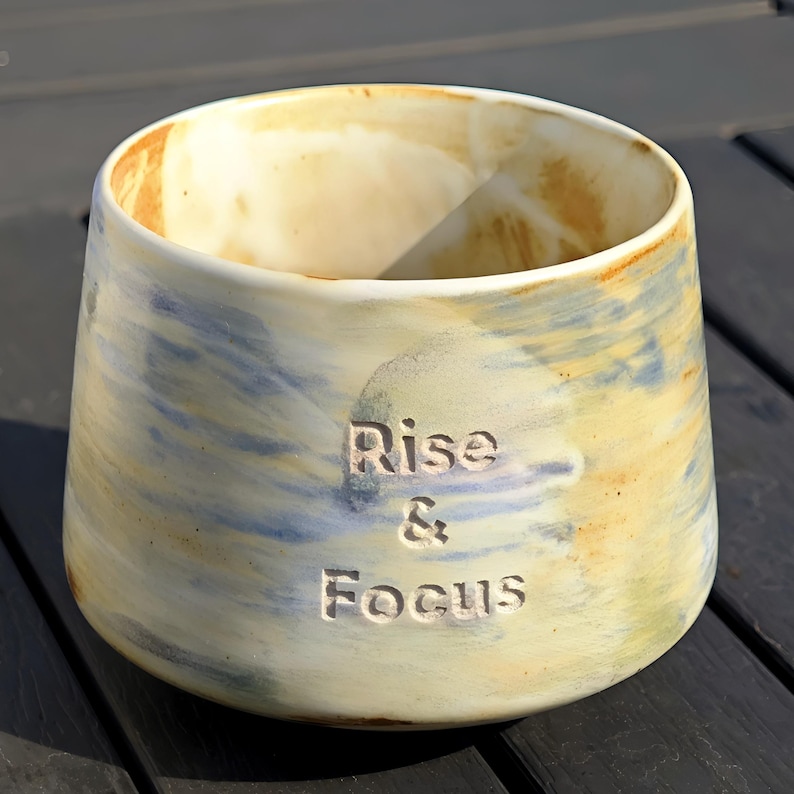 May include: A ceramic mug with a wide opening and a slightly tapered base. The mug is painted with a blend of yellow, blue, and cream colors. The words "Rise & Focus" are carved into the side of the mug.