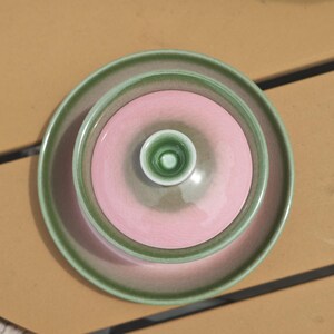 Puede incluir: Un juego de t&eacute; de cer&aacute;mica compuesto por un cuenco y una tapa a juego con un peque&ntilde;o pomo. El conjunto presenta un acabado brillante con un centro rosa suave y un borde verde salvia, creando un dise&ntilde;o circular en capas visto desde arriba.