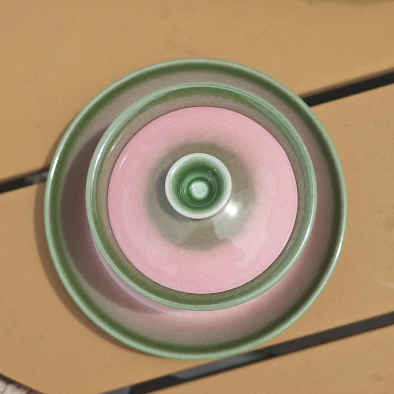 Puede incluir: Un juego de t&eacute; de cer&aacute;mica compuesto por un cuenco y una tapa a juego con un peque&ntilde;o pomo. El conjunto presenta un acabado brillante con un centro rosa suave y un borde verde salvia, creando un dise&ntilde;o circular en capas visto desde arriba.