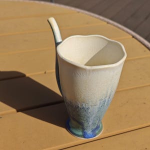 May include: A handmade ceramic mug with a unique handle. The mug features a white top that fades into blue and green hues towards the base. The mug is sitting on a wooden surface.