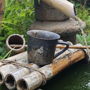 May include: A dark grey ceramic mug with a black handle, resting on bamboo. The mug has a textured surface and a white graphic design. Water flows from a bamboo spout into a small pond, creating a tranquil outdoor scene.