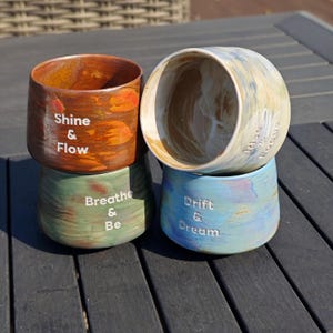 May include: Four stacked ceramic planters with inspirational phrases. The top planter is brown with the words "Shine & Flow". Below it is a green planter with "Breathe & Be". The next planter is blue with "Drift & Dream". The top planter is light blue and white.
