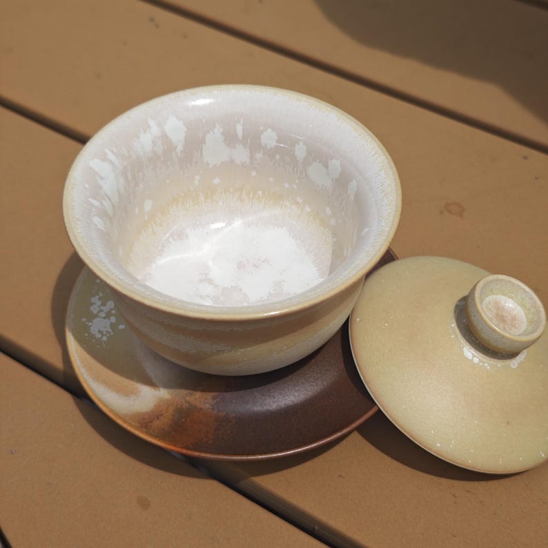 May include: A ceramic tea bowl with a lid and saucer. The bowl features a white interior with a speckled glaze, transitioning to a tan exterior. The saucer is dark brown with a lighter tan rim, and the matching lid is a solid tan color.