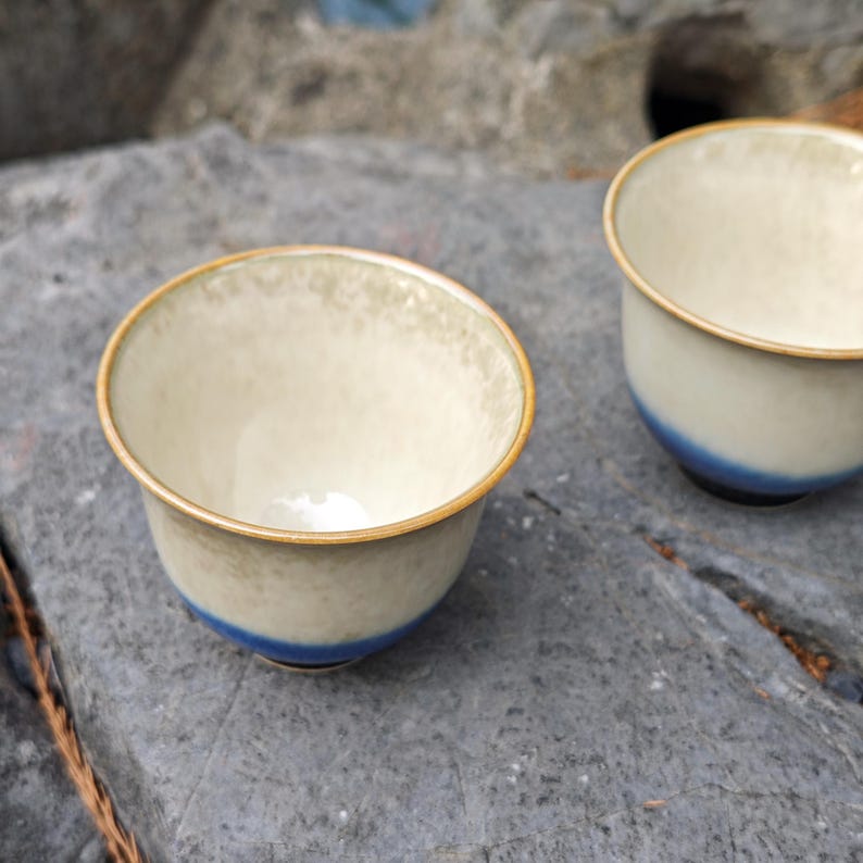 May include: Two ceramic tea cups with a cream-colored interior and a blue band near the base. The rims are outlined in gold. The cups are set on a gray stone surface.