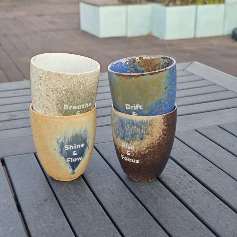 Puede incluir: Cuatro tazas de cer&aacute;mica apiladas, con un esmalte texturizado en tonos beige, azul y marr&oacute;n. Cada taza tiene una palabra inspiradora: "Breathe", "Shine & Flow", "Drift" y "Rise & Focus". Est&aacute;n sobre una superficie de madera gris.