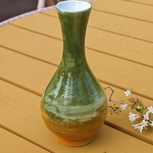 K&ouml;nnte beinhalten: Eine handgefertigte Keramikvase mit schmalem Hals und rundem K&ouml;rper. Die Vase hat einen Farbverlauf, der von einem gelben Sockel zu Gr&uuml;n- und Wei&szlig;t&ouml;nen &uuml;bergeht. Ein kleiner Zweig wei&szlig;er Blumen liegt daneben. Die Vase ist etwa 25 cm hoch.