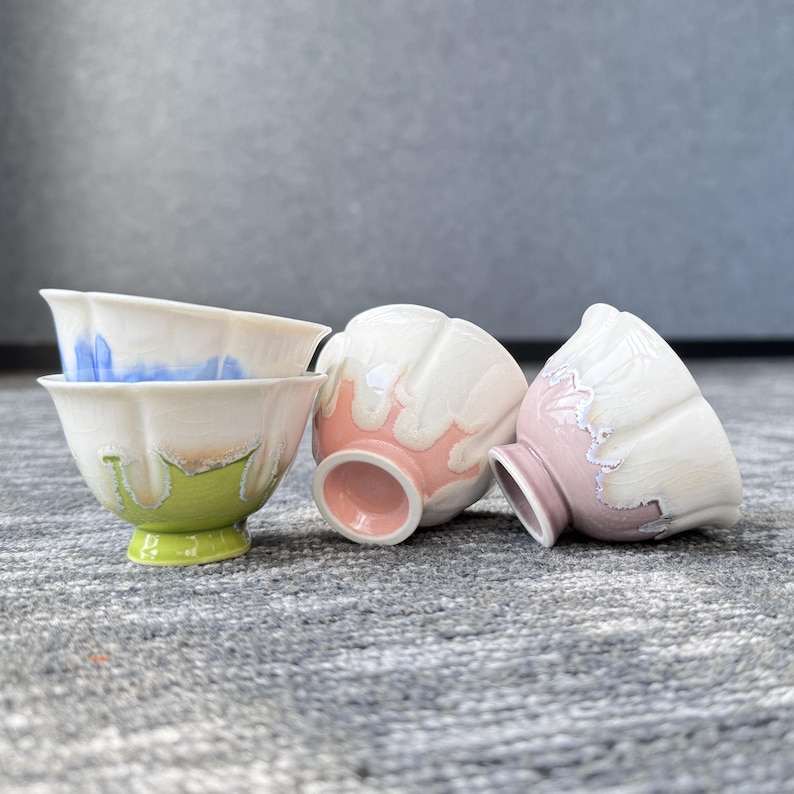 Pode incluir: Quatro ch&aacute;venas de ch&aacute; de cer&acirc;mica com um design &uacute;nico de p&eacute;talas. As ch&aacute;venas apresentam uma base branca com detalhes coloridos em azul, rosa e verde. As ch&aacute;venas est&atilde;o dispostas numa superf&iacute;cie cinzenta.