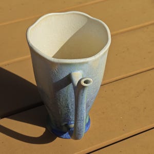 May include: A handmade ceramic pitcher with a unique, irregular rim. The pitcher features a handle with a spout and is painted with a blend of blue, green, and cream colors. The base is a darker blue.