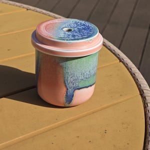 Pu&ograve; includere: Un contenitore in ceramica con coperchio. Il contenitore &egrave; cilindrico con una base rosa, che sfuma in tonalit&agrave; verdi e blu. Il coperchio &egrave; rosa con un centro blu e rosso e un piccolo foro. Il contenitore &egrave; appoggiato su una superficie gialla.