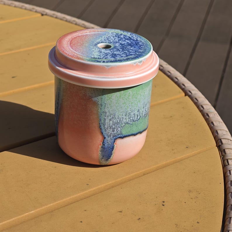Pu&ograve; includere: Un contenitore in ceramica con coperchio. Il contenitore &egrave; cilindrico con una base rosa, che sfuma in tonalit&agrave; verdi e blu. Il coperchio &egrave; rosa con un centro blu e rosso e un piccolo foro. Il contenitore &egrave; appoggiato su una superficie gialla.