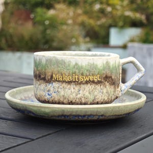May include: A ceramic coffee cup and saucer set with a speckled, textured finish in shades of green, brown, and blue. The cup has a gold inscription that reads "Make it sweet."