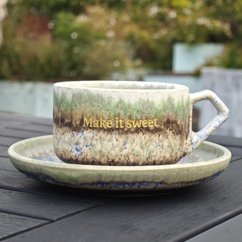 May include: A ceramic coffee cup and saucer set with a speckled, textured finish in shades of green, brown, and blue. The cup has a gold inscription that reads "Make it sweet."