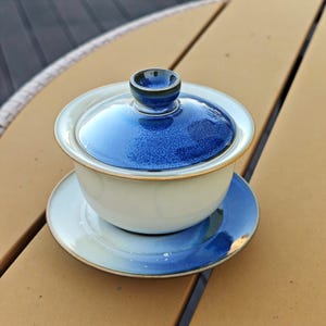 May include: A ceramic gaiwan tea set consisting of a bowl, a lid, and a saucer. The bowl is off-white with a smooth finish. The lid and the saucer feature a deep blue glaze with a textured, speckled appearance and a dark brown rim.