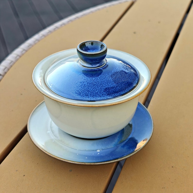 May include: A ceramic gaiwan tea set consisting of a bowl, a lid, and a saucer. The bowl is off-white with a smooth finish. The lid and the saucer feature a deep blue glaze with a textured, speckled appearance and a dark brown rim.