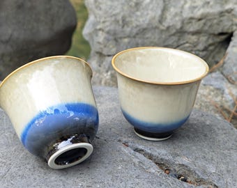 Puede incluir: Dos tazas de té de cerámica con una mitad superior de color crema y una vibrante banda azul en la base. Las tazas tienen un borde dorado y una base negra. Las tazas están colocadas sobre una superficie de piedra gris.