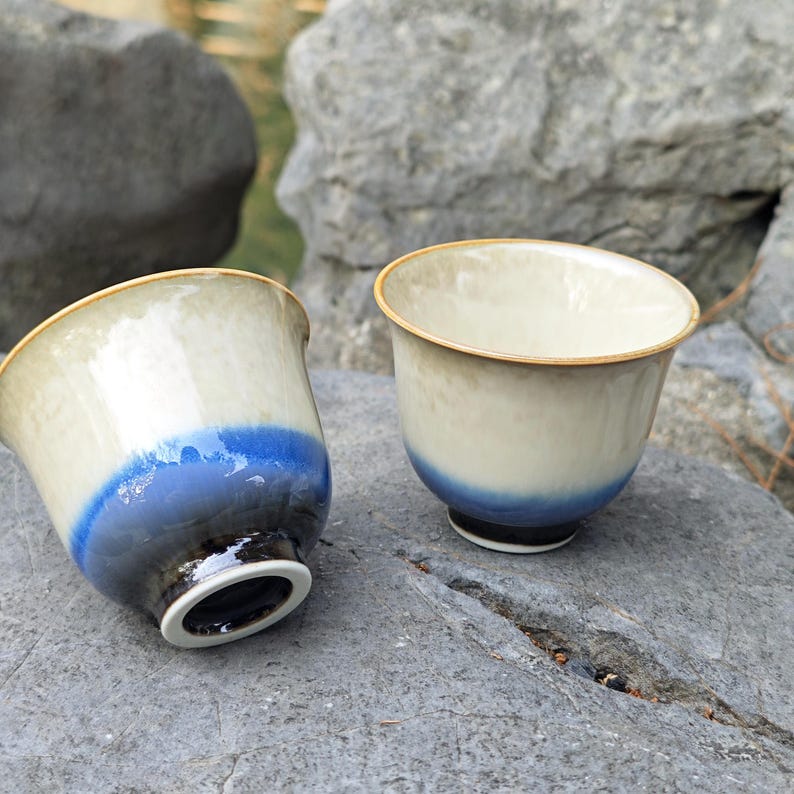 May include: Two ceramic tea cups with a cream-colored upper half and a vibrant blue band at the base. The cups have a gold rim and a black base. The cups are placed on a gray stone surface.