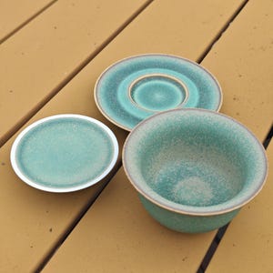 K&ouml;nnte beinhalten: Ein Keramikset bestehend aus einer Schale, einer Untertasse und einem kleinen Teller, alle mit einer gesprenkelten t&uuml;rkis-gr&uuml;nen Glasur und einem glatten, hellen Rand. Die Teile weisen eine gleichm&auml;&szlig;ige, gesprenkelte Textur und ein abgerundetes Design auf.