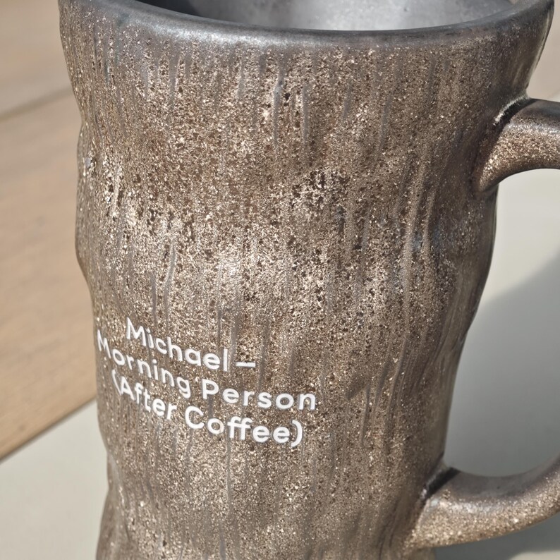 Puede incluir: Una taza de cer&aacute;mica de color marr&oacute;n oscuro con una superficie texturizada que recuerda a la corteza de un &aacute;rbol. La taza tiene un asa en el lado derecho y un texto impreso en blanco que dice: Michael - Morning Person (After Coffee).