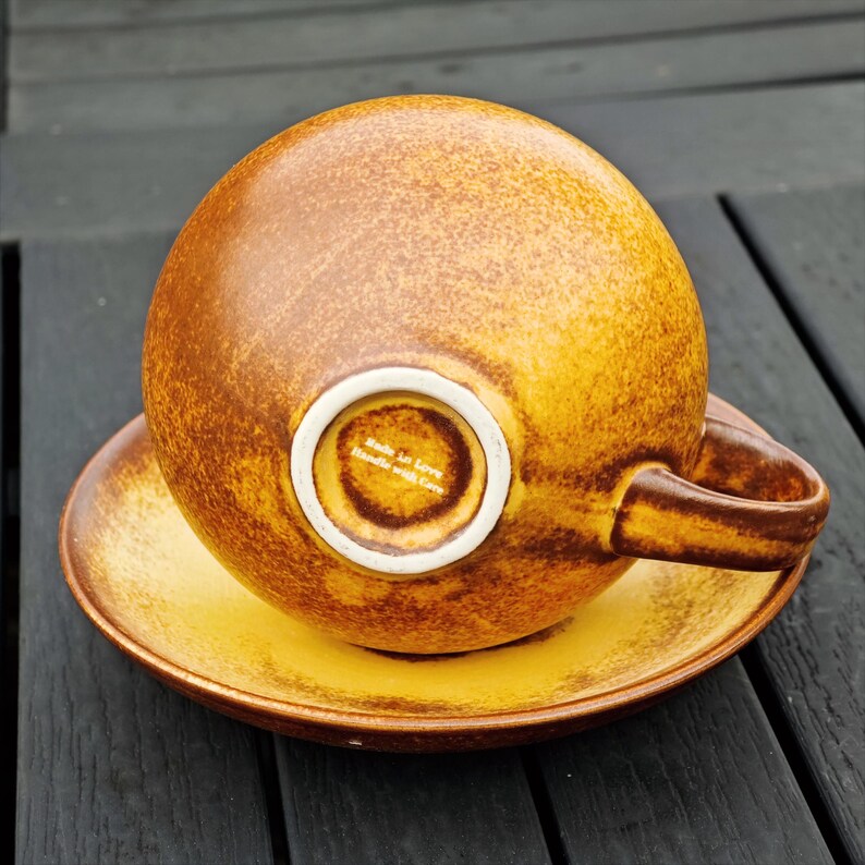 May include: A brown and yellow ceramic teacup and saucer set. The cup is upside down, revealing a white interior with text. The saucer is round and matches the cup's color scheme. The set has a textured, speckled appearance.