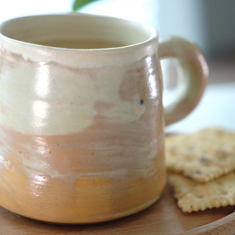 May include: A handmade ceramic mug with a cream, peach, and tan glaze. The mug has a rounded shape with a large handle. Two crackers are on a wooden tray next to the mug. The mug is filled with a light-colored liquid.