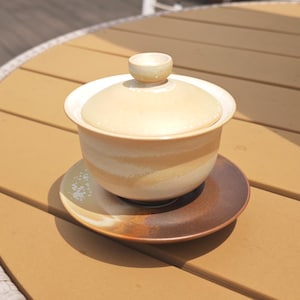 May include: A ceramic gaiwan tea set featuring a lidded bowl with a cream and beige gradient glaze, resting on a matching saucer that transitions from a light cream to a deep, dark brown tone with subtle white speckled accents on the rim.