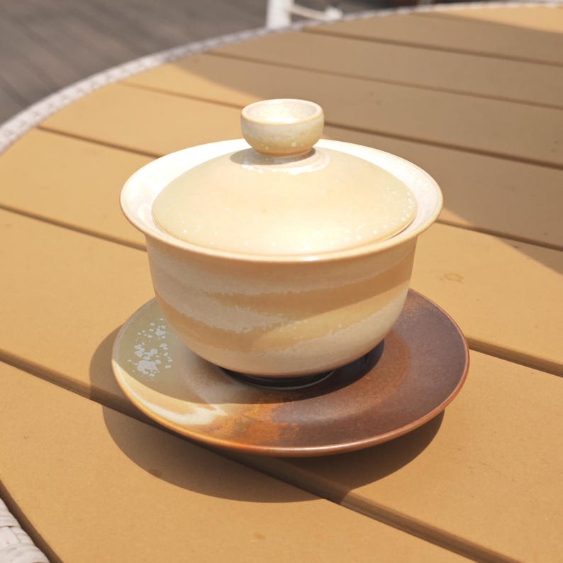 May include: A ceramic gaiwan tea set featuring a lidded bowl with a cream and beige gradient glaze, resting on a matching saucer that transitions from a light cream to a deep, dark brown tone with subtle white speckled accents on the rim.