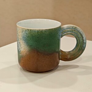 K&ouml;nnte beinhalten: Eine Keramik-Tasse mit einem einzigartigen Griffdesign. Die Tasse hat einen Farbverlauf aus Braun, Gr&uuml;n und Blau mit einem wei&szlig;en Innenraum. Der Griff ist kreisf&ouml;rmig mit einer strukturierten Oberfl&auml;che.