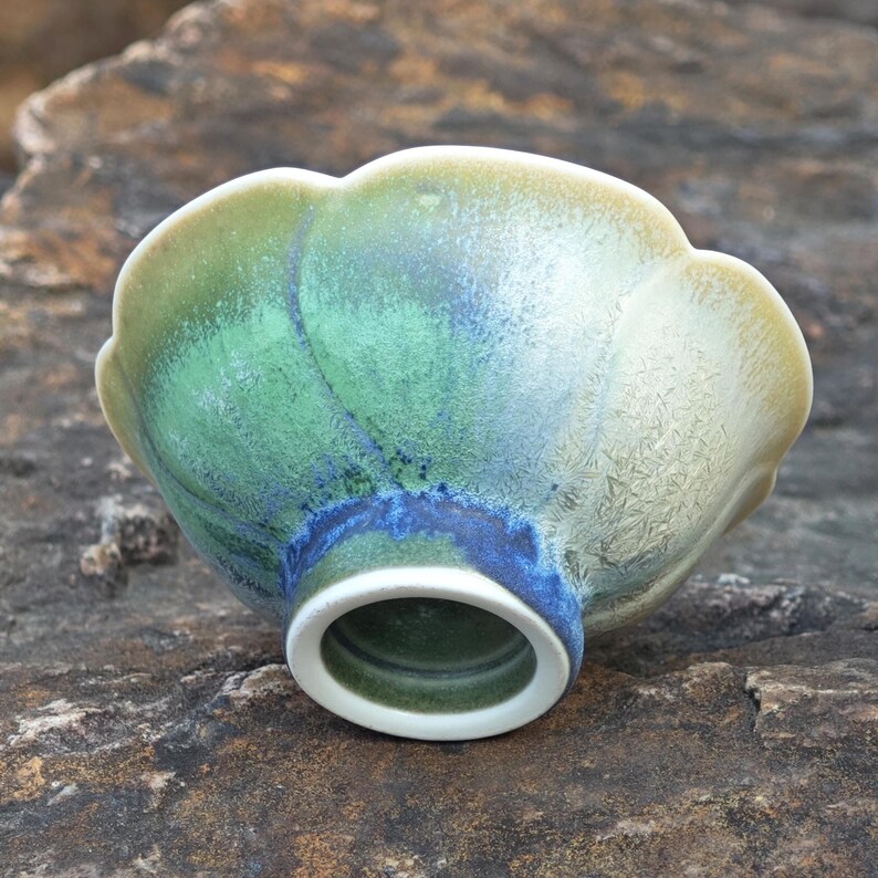 May include: A ceramic bowl with a scalloped rim, showcasing a blend of green, blue, and cream hues. The bowl's exterior features a textured, glazed finish, and a white rim around the base. The bowl is resting on a rough, stone surface.