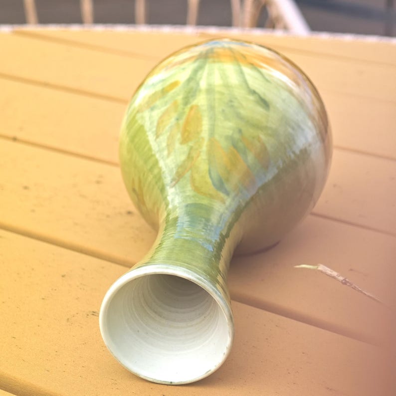 K&ouml;nnte beinhalten: Eine handgefertigte Keramikvase mit bauchigem K&ouml;rper und langem Hals. Die Vase ist mit gr&uuml;nen, gelben und blauen Bl&auml;ttermustern bemalt. Das Innere der Vase ist wei&szlig;. Die Vase steht auf einer Holzoberfl&auml;che.