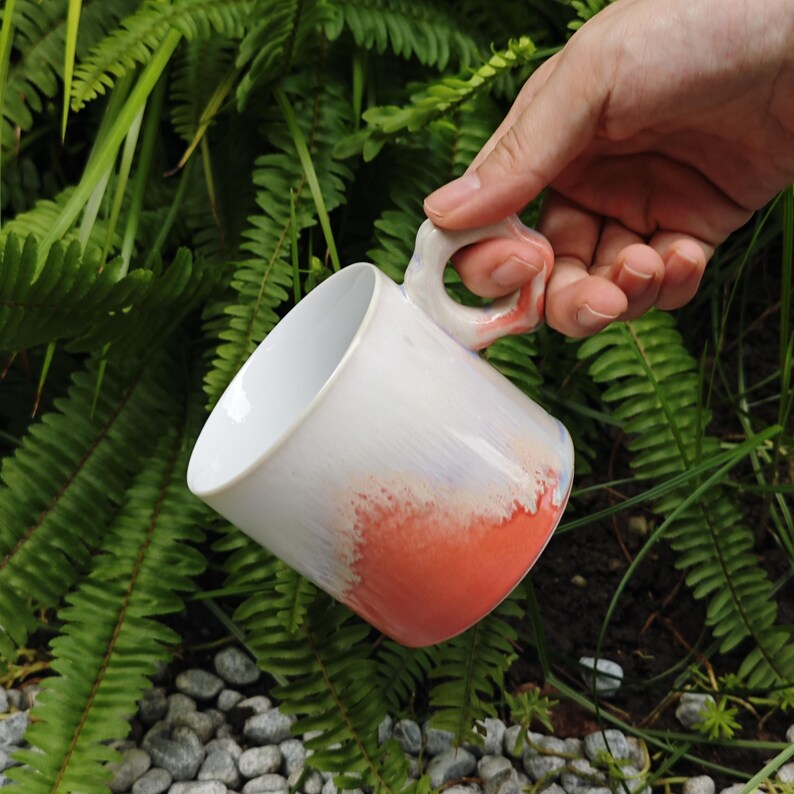 May include: White ceramic mug with a unique handle, featuring a gradient of colors from white to orange. The mug is held against a backdrop of green foliage, showcasing its design and shape.