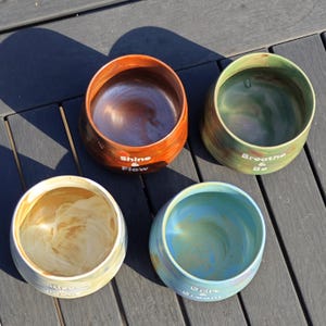 May include: Four handcrafted ceramic bowls in shades of brown, green, blue, and cream. Each bowl has a rounded shape and features a unique color gradient. The bowls have inspirational text: "Shine & Flow", "Breathe & Be", and "Spice & Dream".