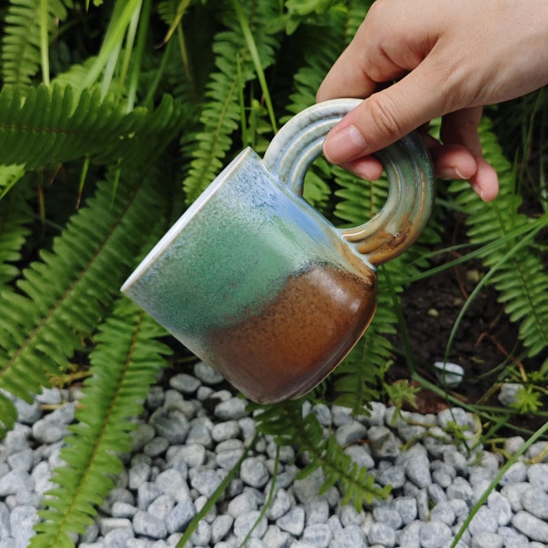 K&ouml;nnte beinhalten: Eine handgefertigte Keramik-Tasse mit einem einzigartigen Griffdesign. Die Tasse weist einen Farbverlauf auf, der Blau, Gr&uuml;n und Braun umfasst. Die Tasse wird vor einem Hintergrund aus gr&uuml;nem Laub und kleinen grauen Steinen gehalten.