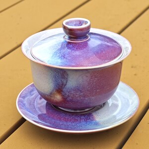 May include: A purple and blue ceramic gaiwan tea set with a matching lid and saucer. The tea set has a glossy finish and is sitting on a wooden surface. The gaiwan is a traditional Chinese tea brewing vessel.