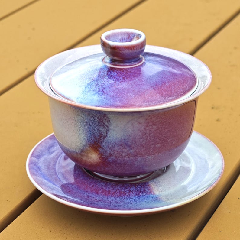 May include: A purple and blue ceramic gaiwan tea set with a matching lid and saucer. The tea set has a glossy finish and is sitting on a wooden surface. The gaiwan is a traditional Chinese tea brewing vessel.