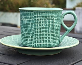 Könnte beinhalten: Ein türkisfarbenes Keramik-Tee-Tassen- und Untertassen-Set. Die Tasse hat einen einzigartigen Henkel und ein strukturiertes Webmuster. Die Untertasse ist farblich passend türkis mit einem hellgrünen Rand. Für Tee oder Kaffee.