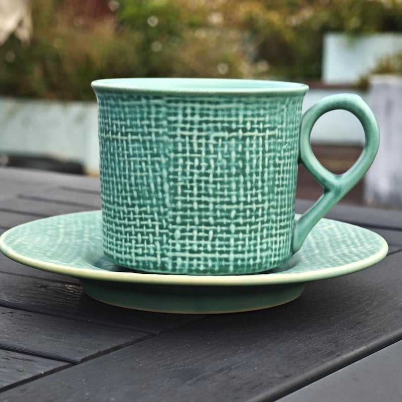 May include: A teal ceramic teacup and saucer set. The cup has a unique handle and a textured woven pattern. The saucer is a matching teal color with a lighter green rim. The set is designed for serving tea or coffee.
