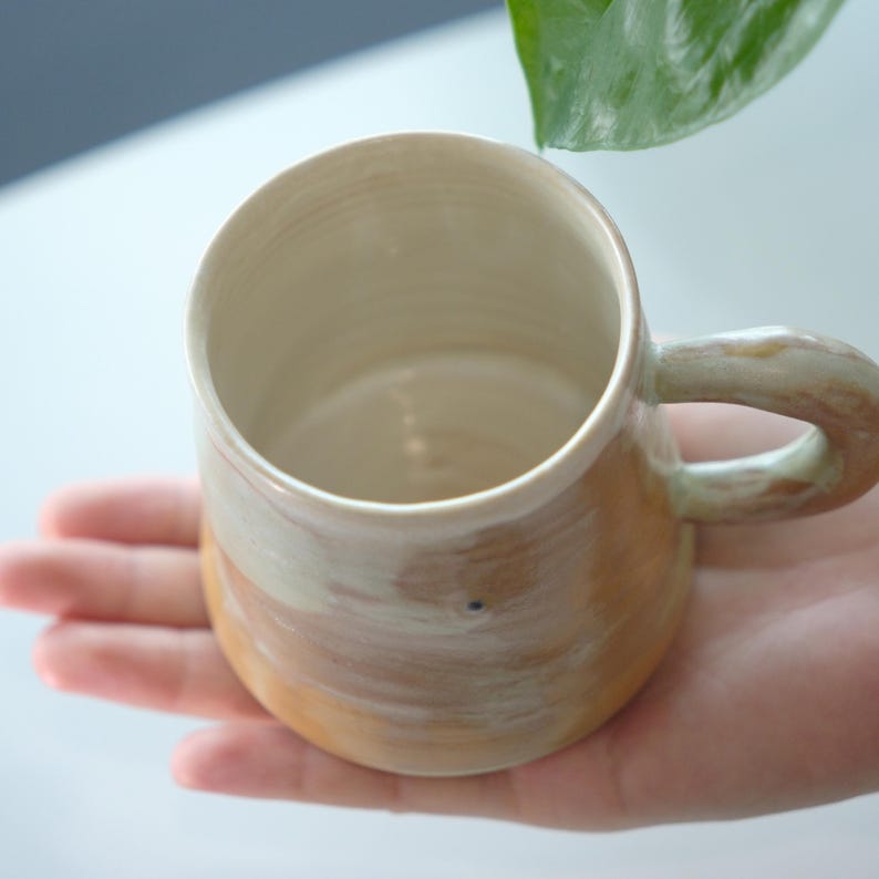 May include: A handmade ceramic mug with a rounded body and a curved handle. The mug features a blend of colors, including cream, tan, and light brown, with a textured, organic appearance. The interior is a solid cream color.