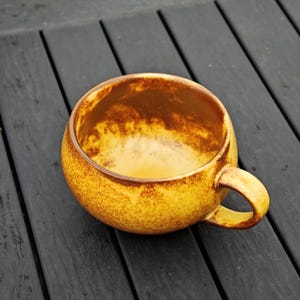 May include: A round, yellow ceramic mug with a brown interior and handle. The mug has a textured surface and sits on a dark wooden surface. The mug is empty and ready to be filled with a warm beverage.