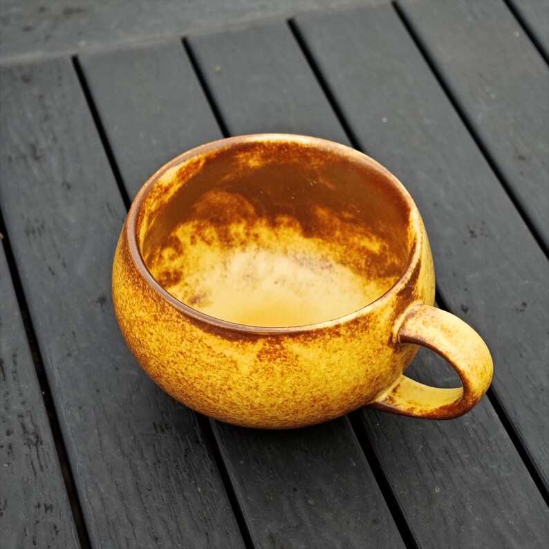 May include: A round, yellow ceramic mug with a brown interior and handle. The mug has a textured surface and sits on a dark wooden surface. The mug is empty and ready to be filled with a warm beverage.