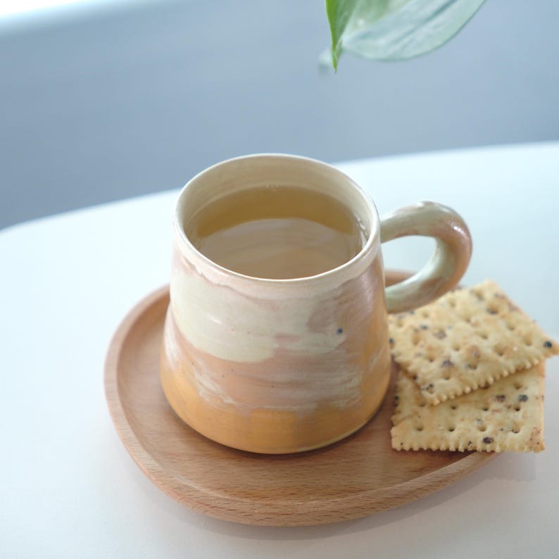 May include: A handmade ceramic mug with a beige and orange glaze, filled with a light-colored beverage, rests on a small wooden plate. Two crackers with seeds are placed next to the mug. The mug has a rounded handle.